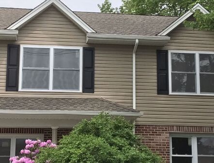 Suburban house facade with beige siding, black shutters, brick lower level, and flowering shrubs