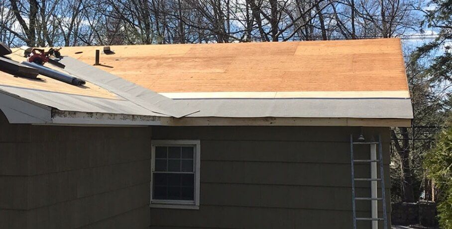 Partially re-roofed house with exposed plywood roof and gray siding under trees.