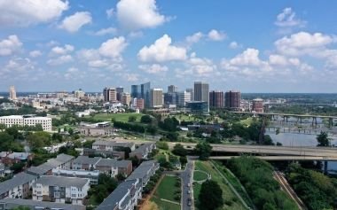 City skyline with modern buildings, green parkland, and a river under a bright blue sky with clouds