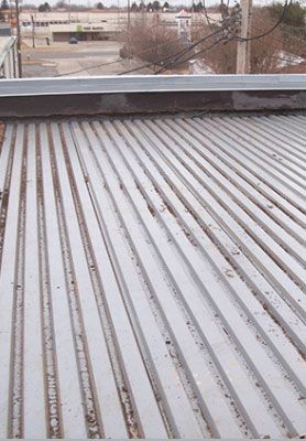 Snow-dusted corrugated metal roof with a low parapet and bare trees in the distance
