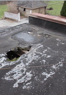 Damaged flat rooftop with a large hole and peeling, weathered black roofing material.