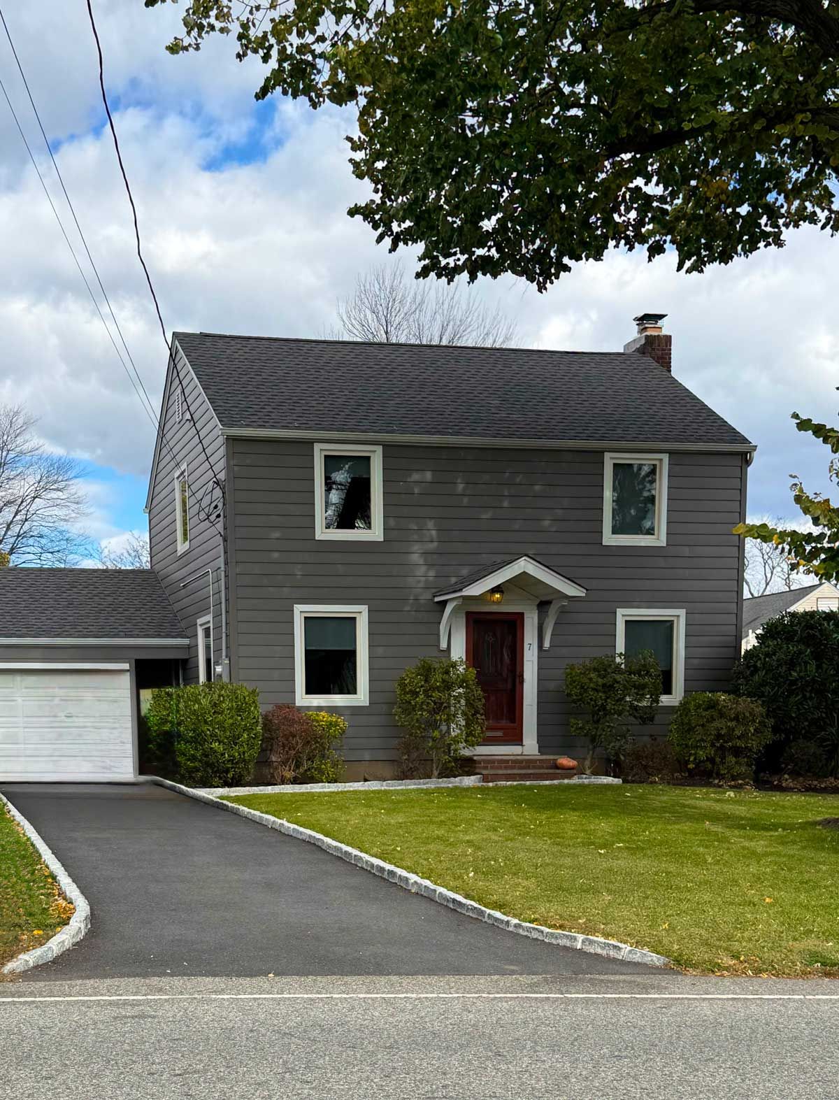 Gray two-story house with white trim, red front door, driveway, and green lawn under cloudy sky