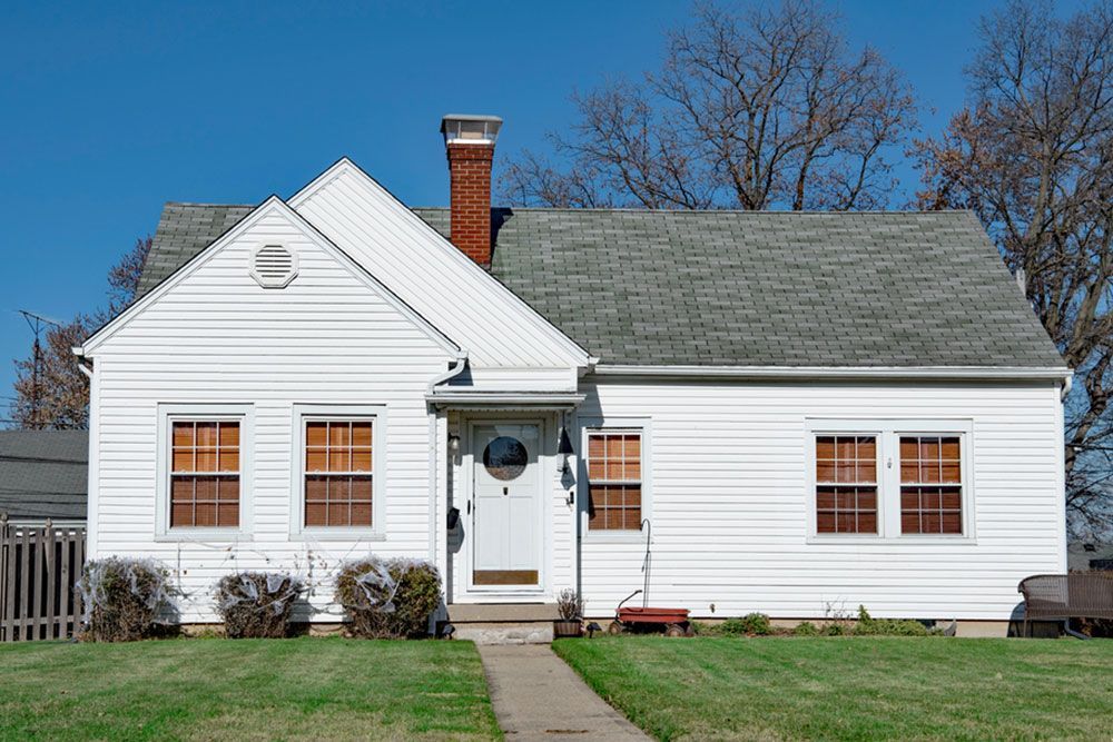 Small white house with a gray roof, brick chimney, and front walkway on a sunny day