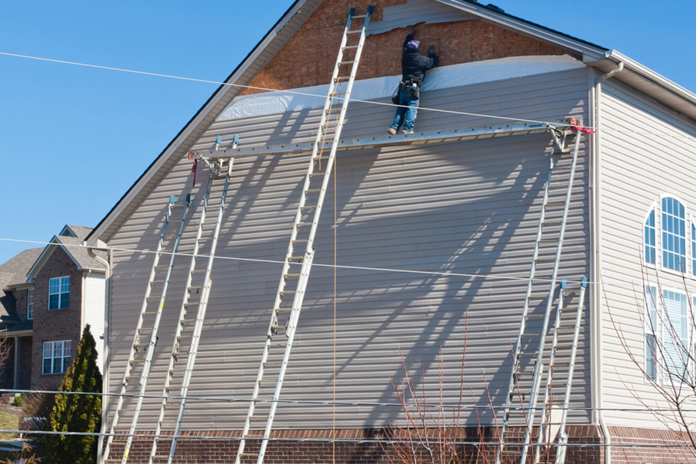 Worker on ladder repairing a brick house roof, with another ladder leaning against the wall.
