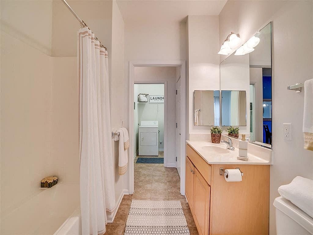Bright bathroom with shower curtain, vanity sink, mirror, and open doorway to another room.