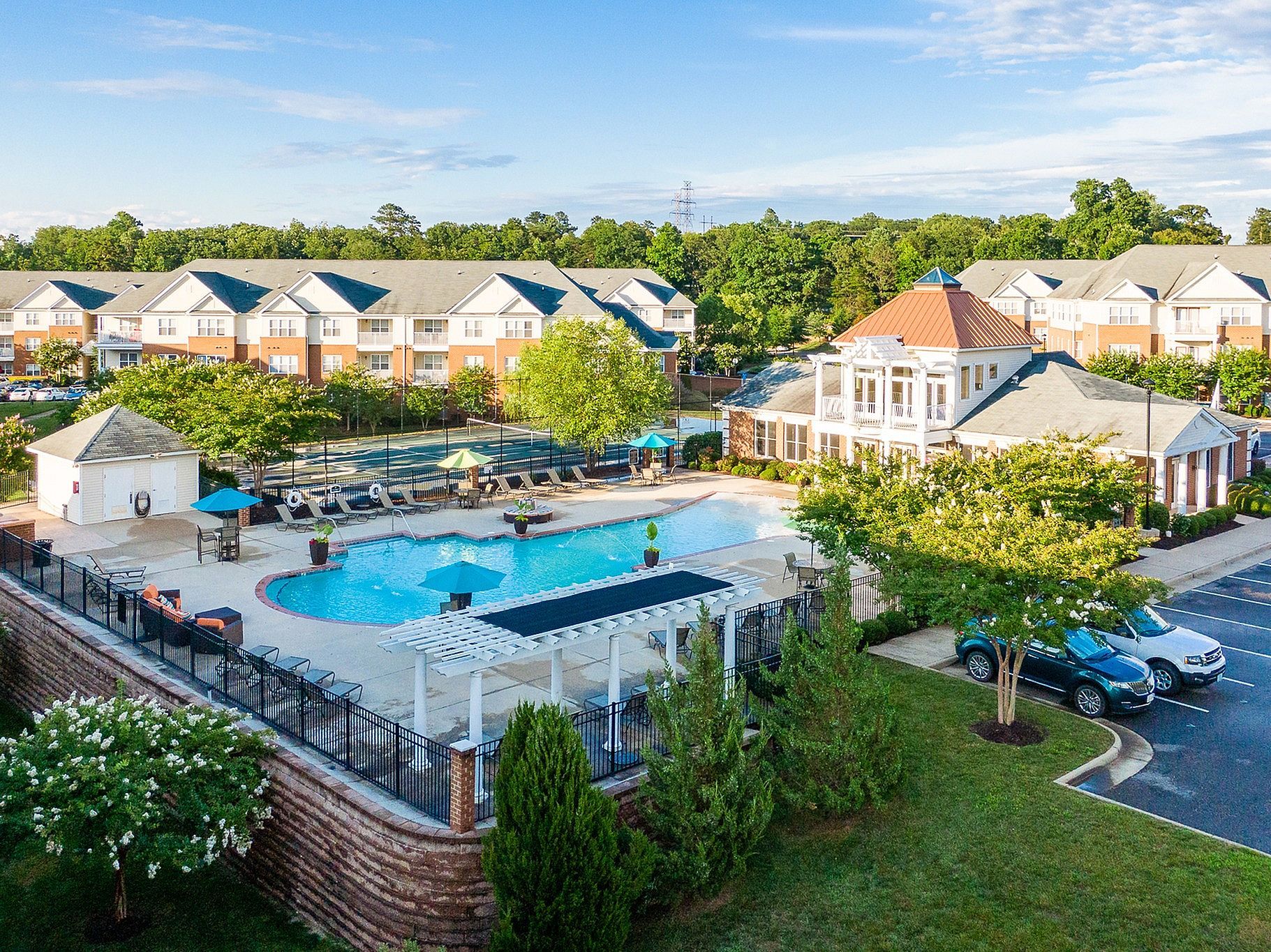Aerial view of a resort pool with surrounding townhouses, trees, and parked cars under a clear sky