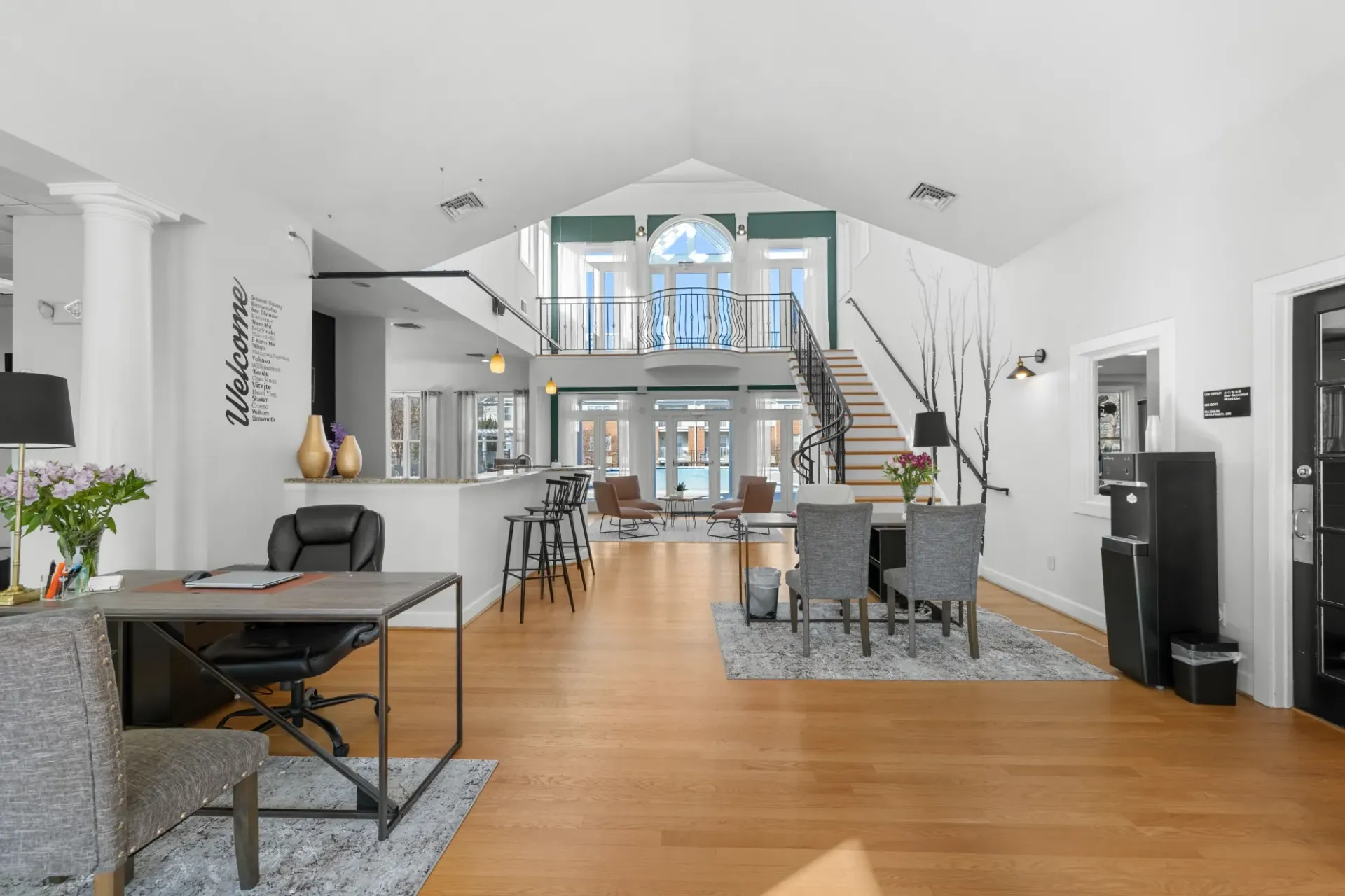 Bright open-concept living room and dining area with wood floors, high ceiling, staircase, and large windows