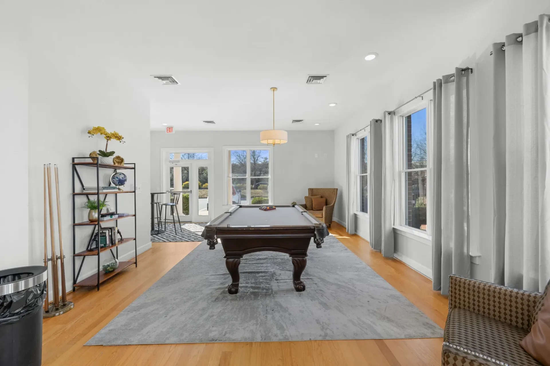Bright living room with a pool table, gray rug, wood floors, and large windows overlooking trees