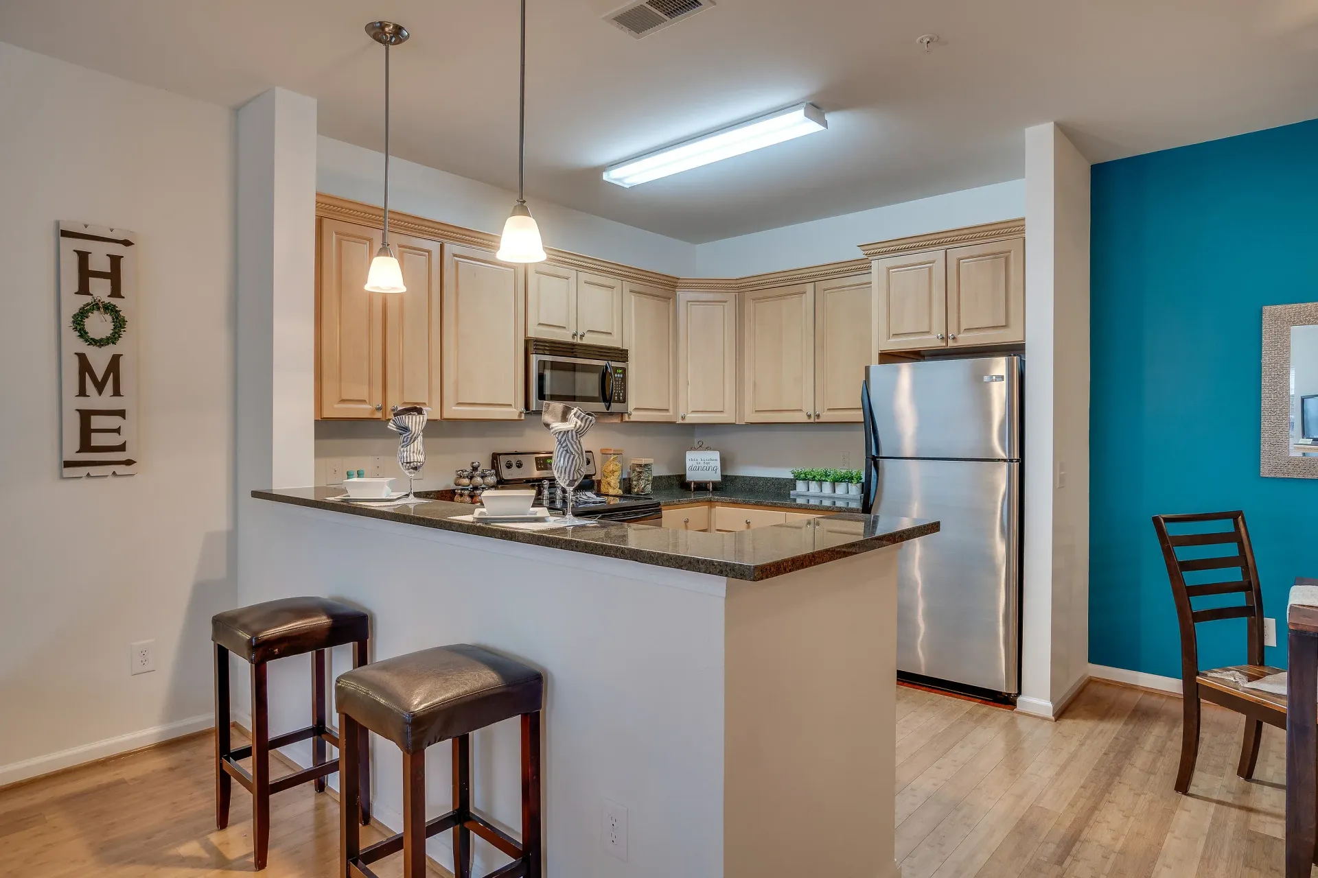 Bright kitchen with granite island, stainless fridge, pendant lights, and two stools; teal dining nook visible