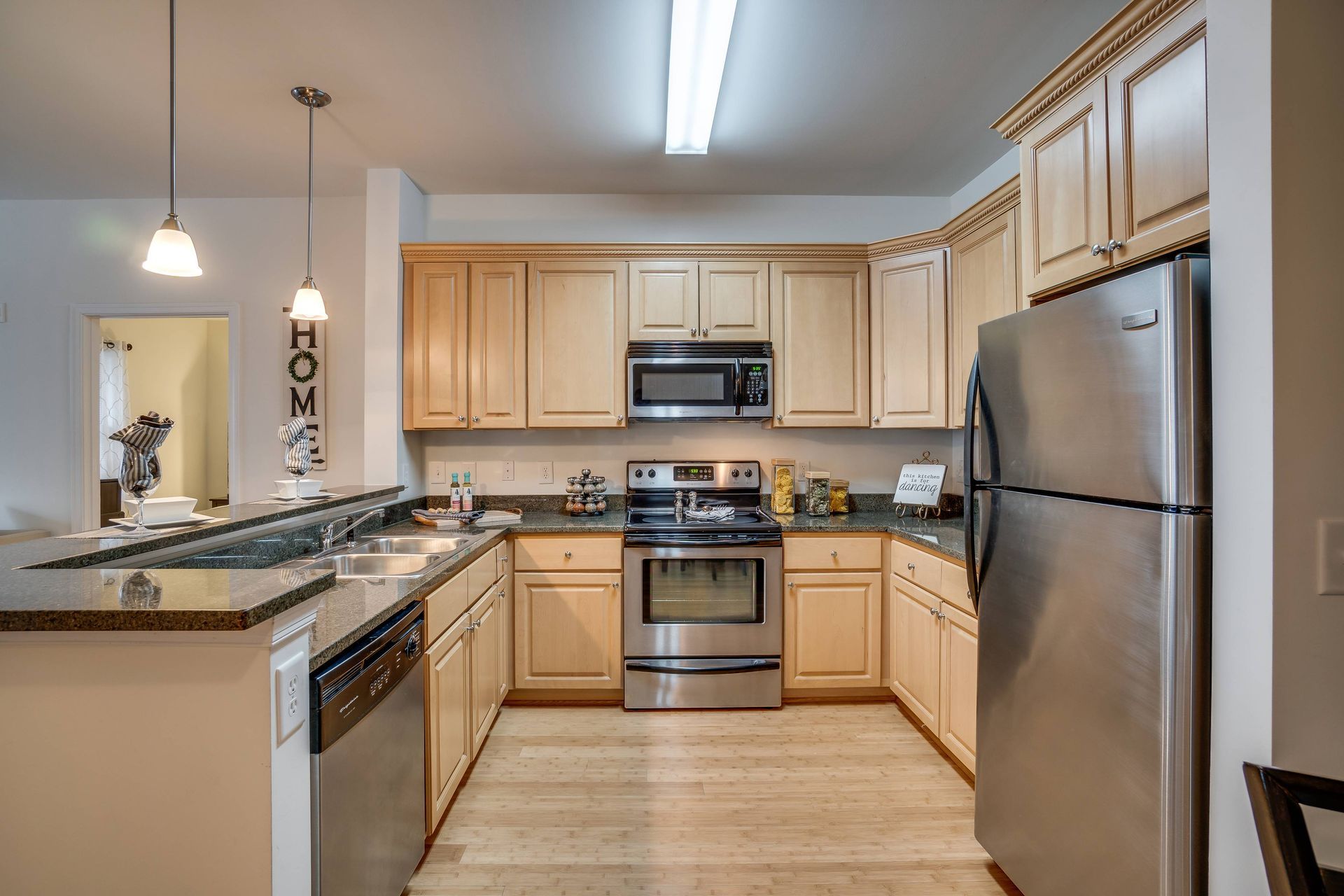 Modern kitchen with light wood cabinets, stainless steel appliances, dark countertops, and hanging pendant lights.