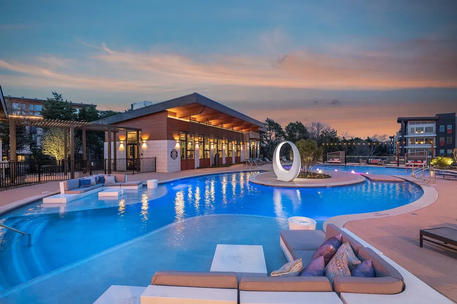 Modern swimming pool and lounge area at dusk with a clubhouse in the background.