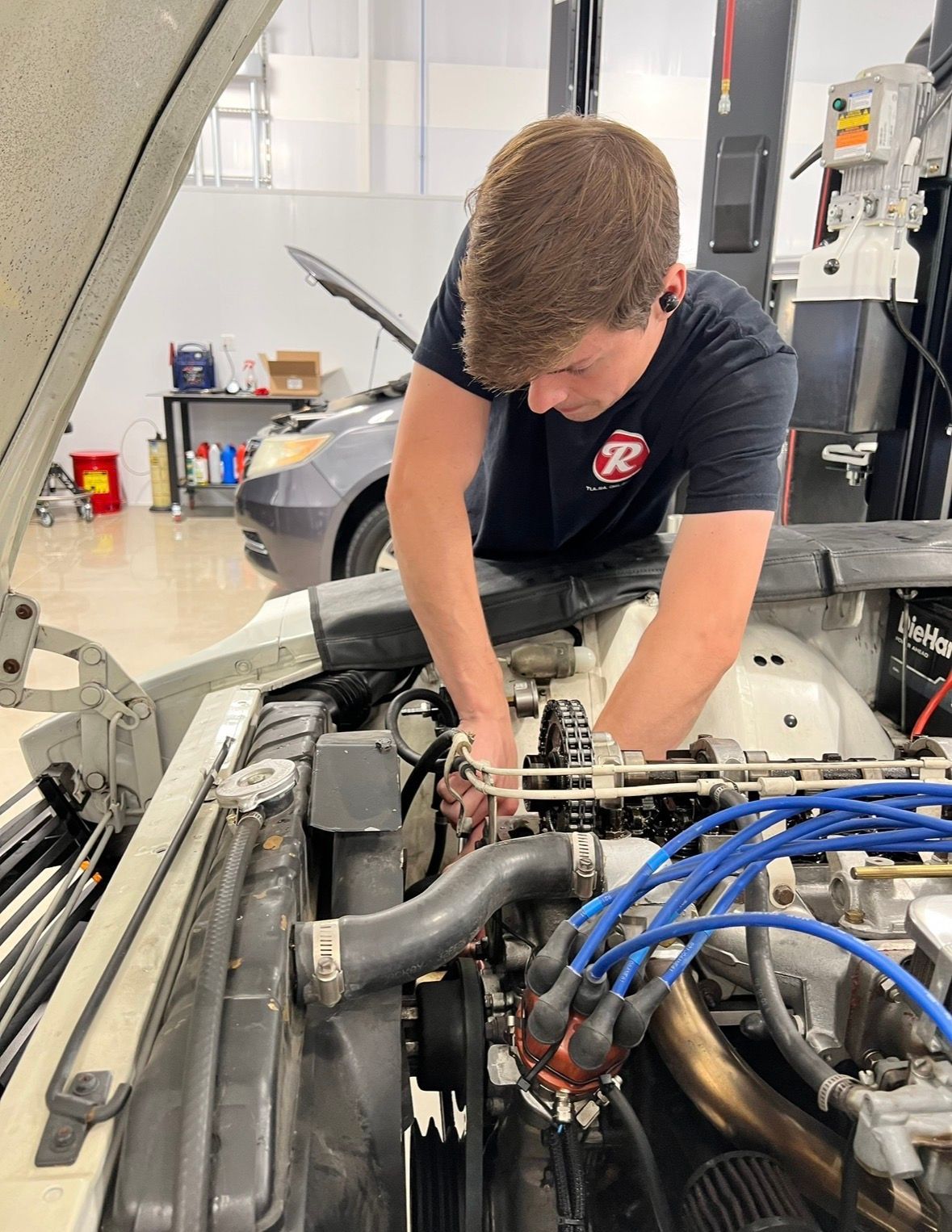 Mechanic working on a car engine in a garage, with blue hoses and open hood. | Reeder's Auto & Tire