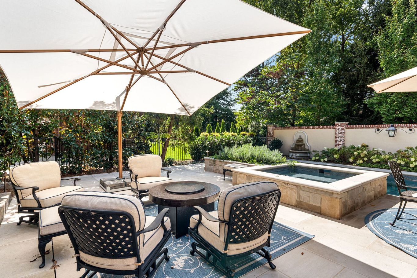 A patio area featuring a white umbrella, fire pit, outdoor furniture, and a stone-encased spa surrounded by greenery.