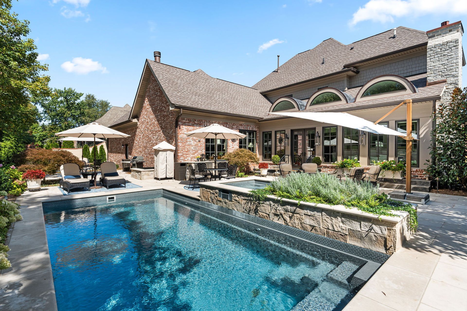 A stone house with a backyard featuring a rectangular pool, a hot tub, stone patio, and patio furniture under umbrellas.