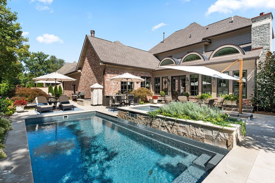 A backyard swimming pool and hot tub next to a large stone house with a patio and umbrellas under a sunny sky.