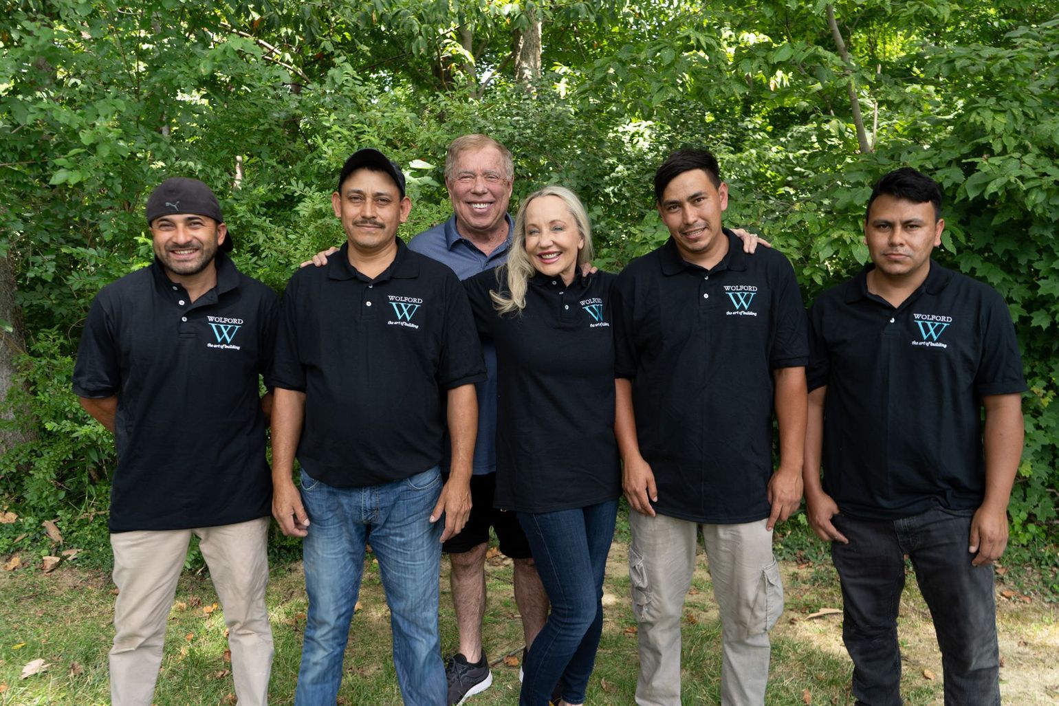 A group of six people stand in front of trees, wearing matching black company polo shirts, posing for a group photo.