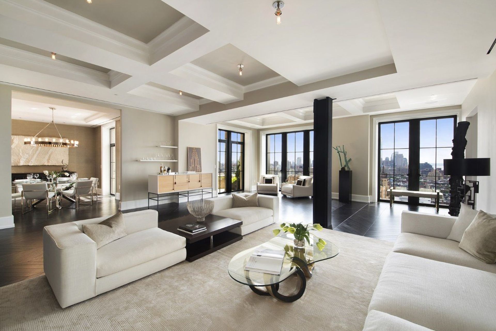A spacious, modern living room with beige sofas, a glass coffee table, dark wood floors, and large windows.