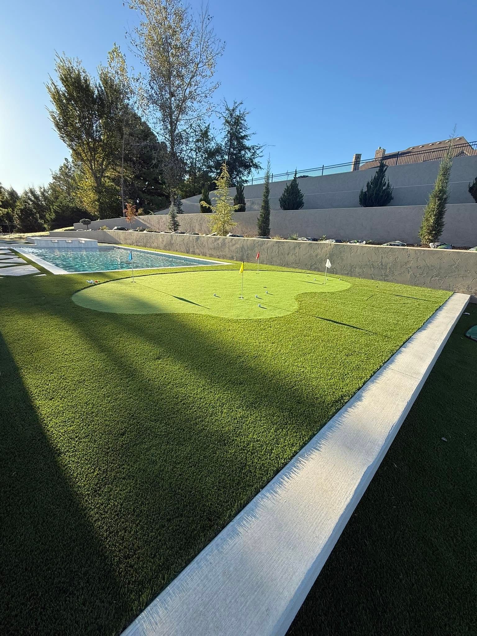 Sunny backyard with green lawn, small pool, trees, and a gray wall under a clear blue sky