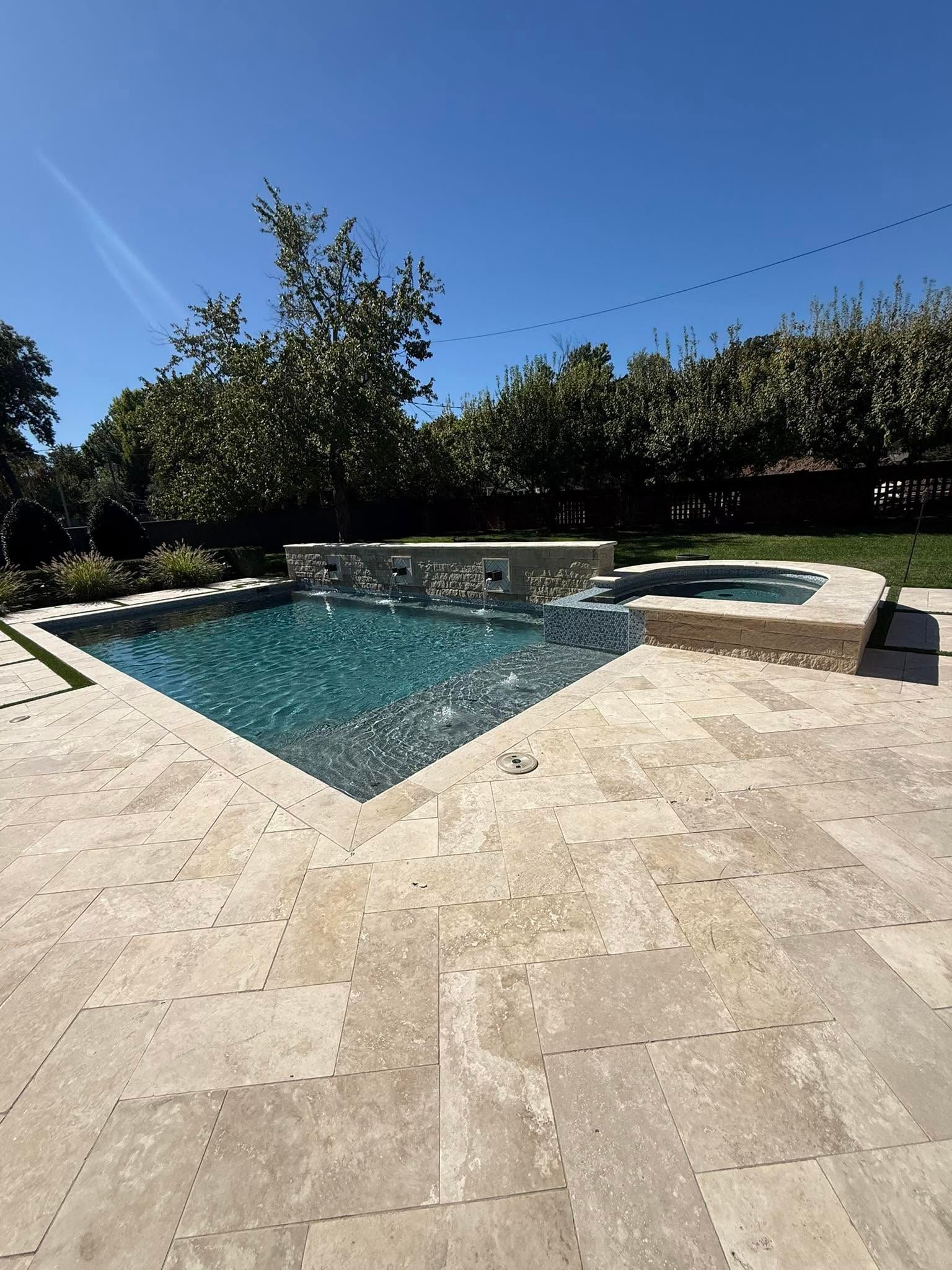 Backyard pool with stone patio, blue water, and green hedges under a clear sky