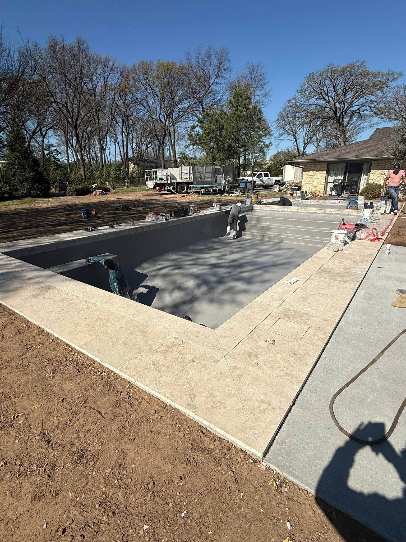 Backyard swimming pool under construction with workers, concrete deck, and trees in the background