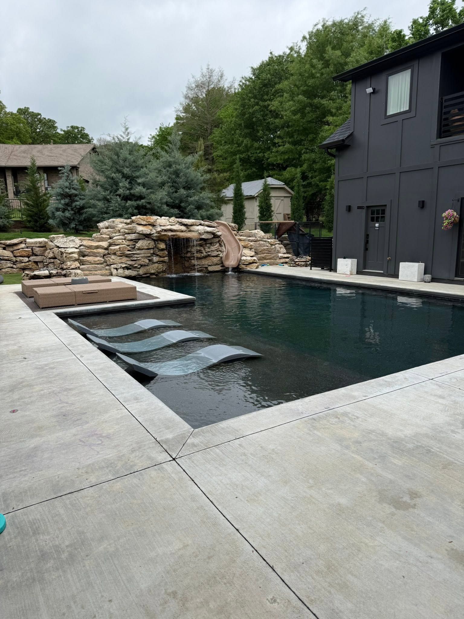 Backyard pool with stone waterfall, concrete patio, and modern black house exterior