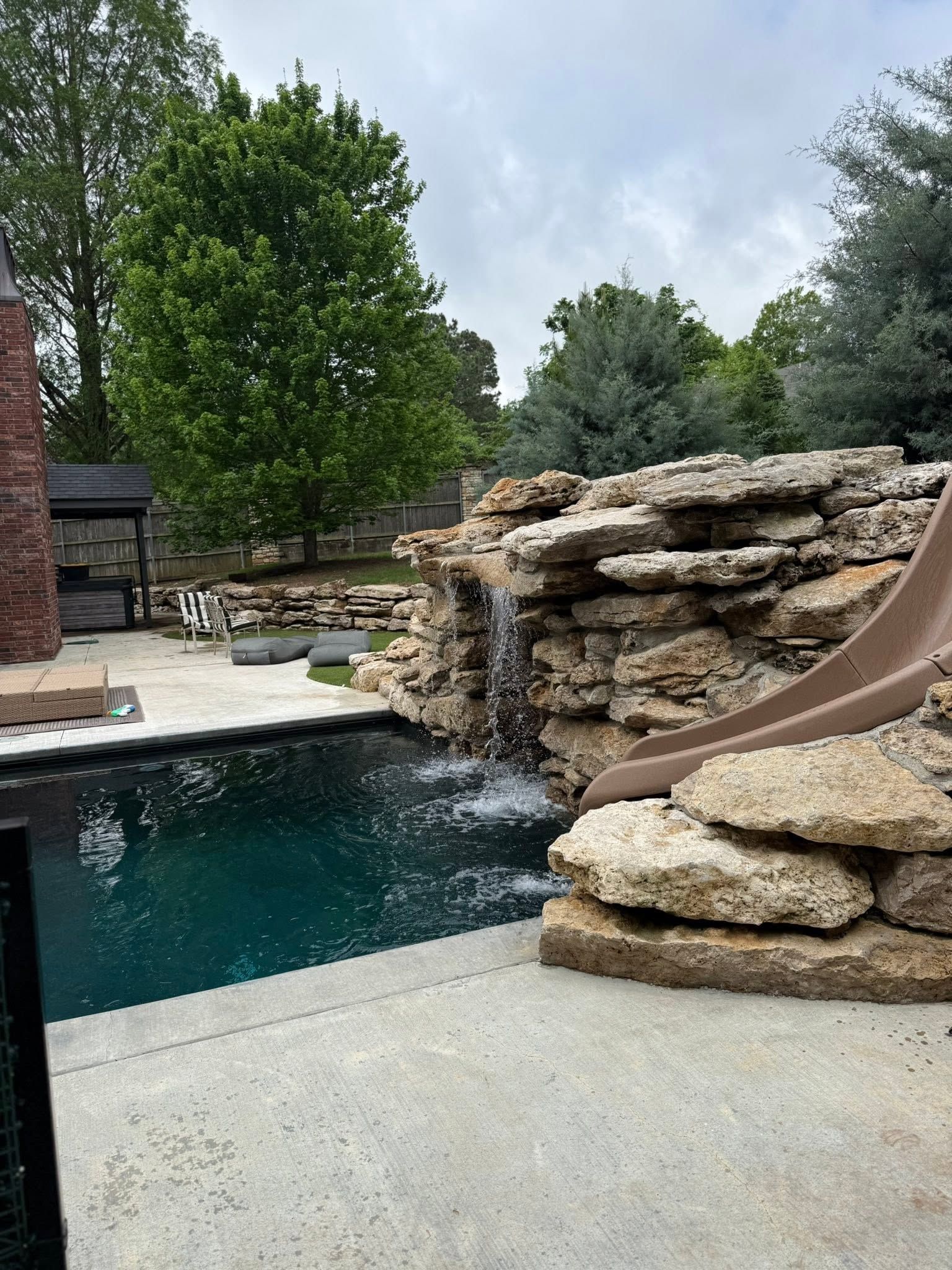 Backyard pool with rock waterfall and slide beside a concrete deck and trees