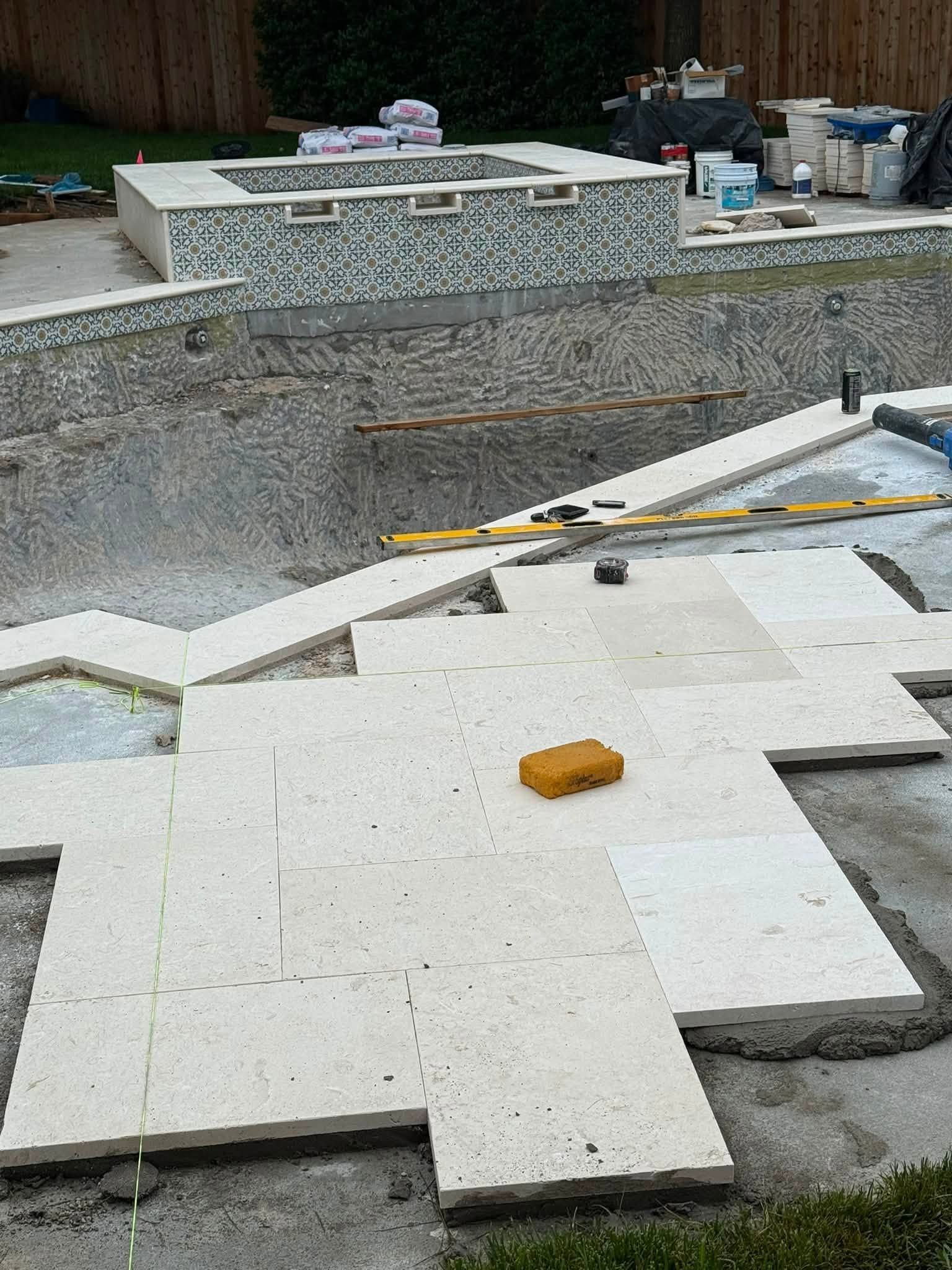 Tiled pool deck under construction with white stone slabs, tools, and a gray stone wall.