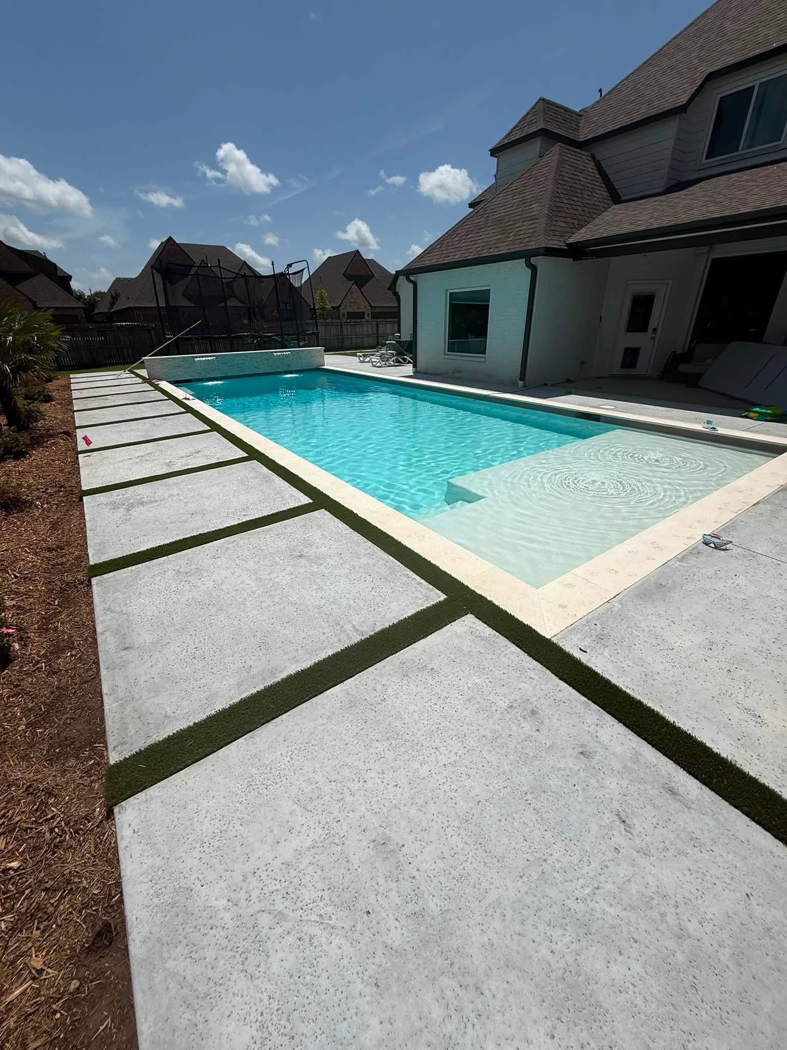 Backyard swimming pool beside a modern house, with white stepping stones and blue sky overhead.
