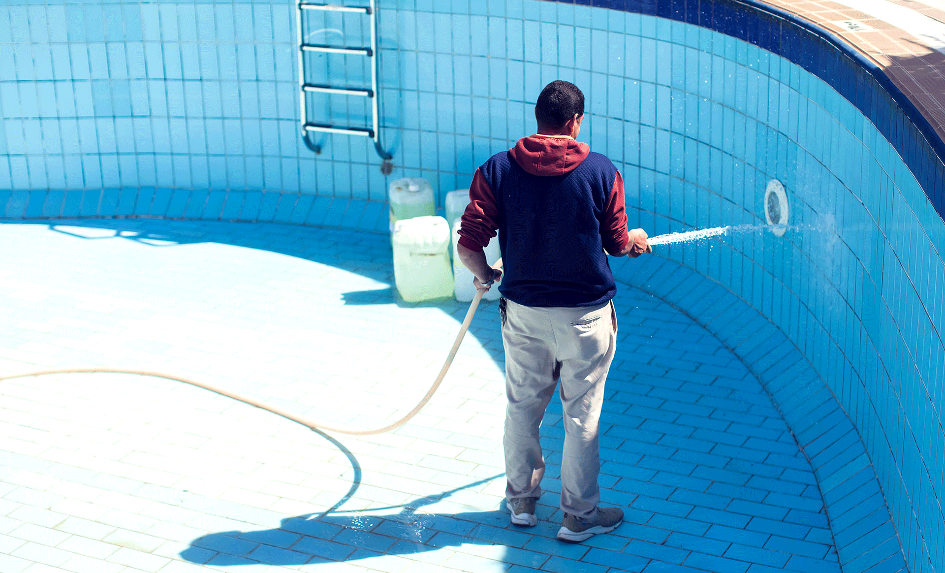 Person sprays the inside of a blue empty pool with a hose and bucket