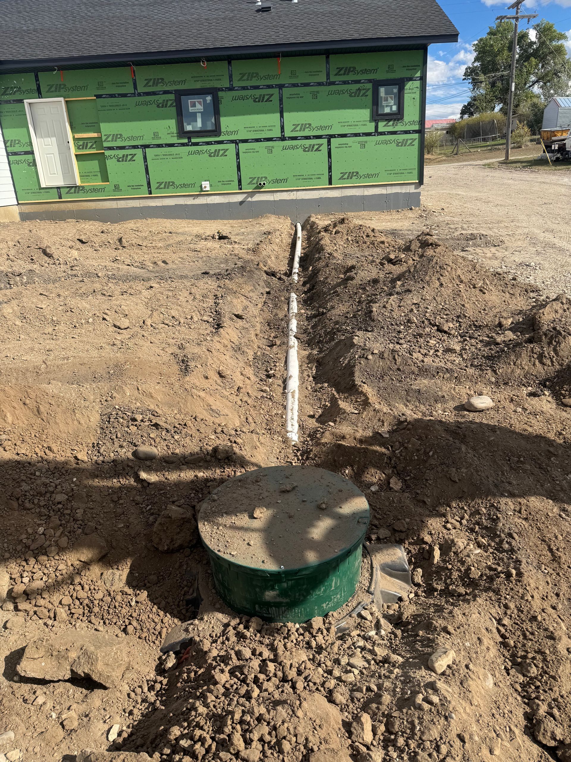 Green building with a black septic tank lid and white pipe in a dirt excavation site.