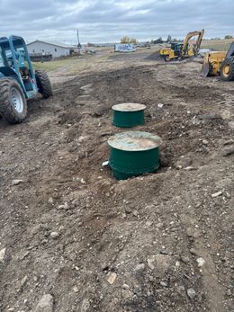 Construction site with two green utility boxes in a muddy dirt lot and heavy equipment in the background