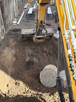 Excavator digging a trench beside a yellow wall, with soil piled around a large tire in the pit