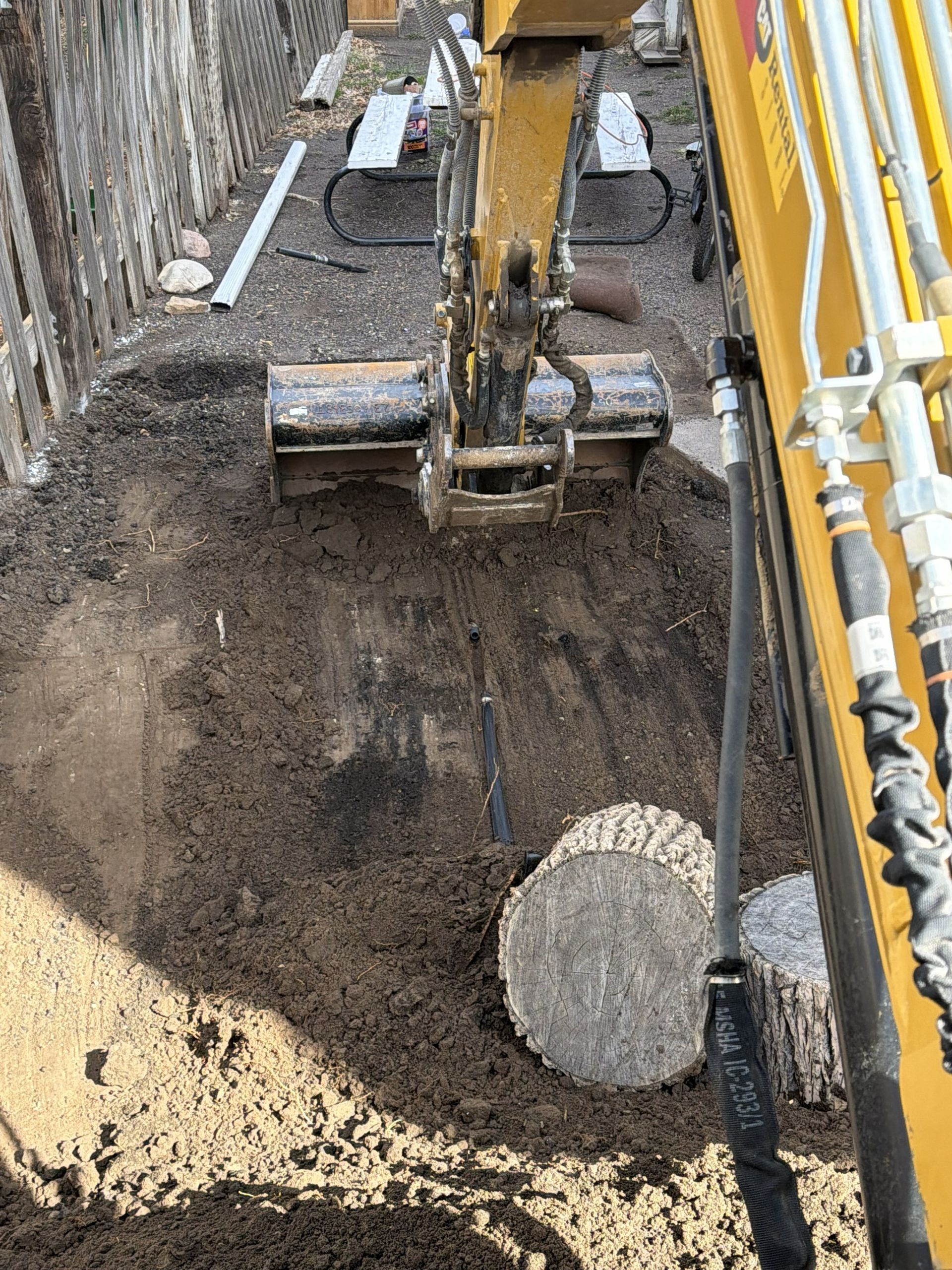 Excavator digging a deep trench beside concrete walls, with dirt and a large pipe section visible.
