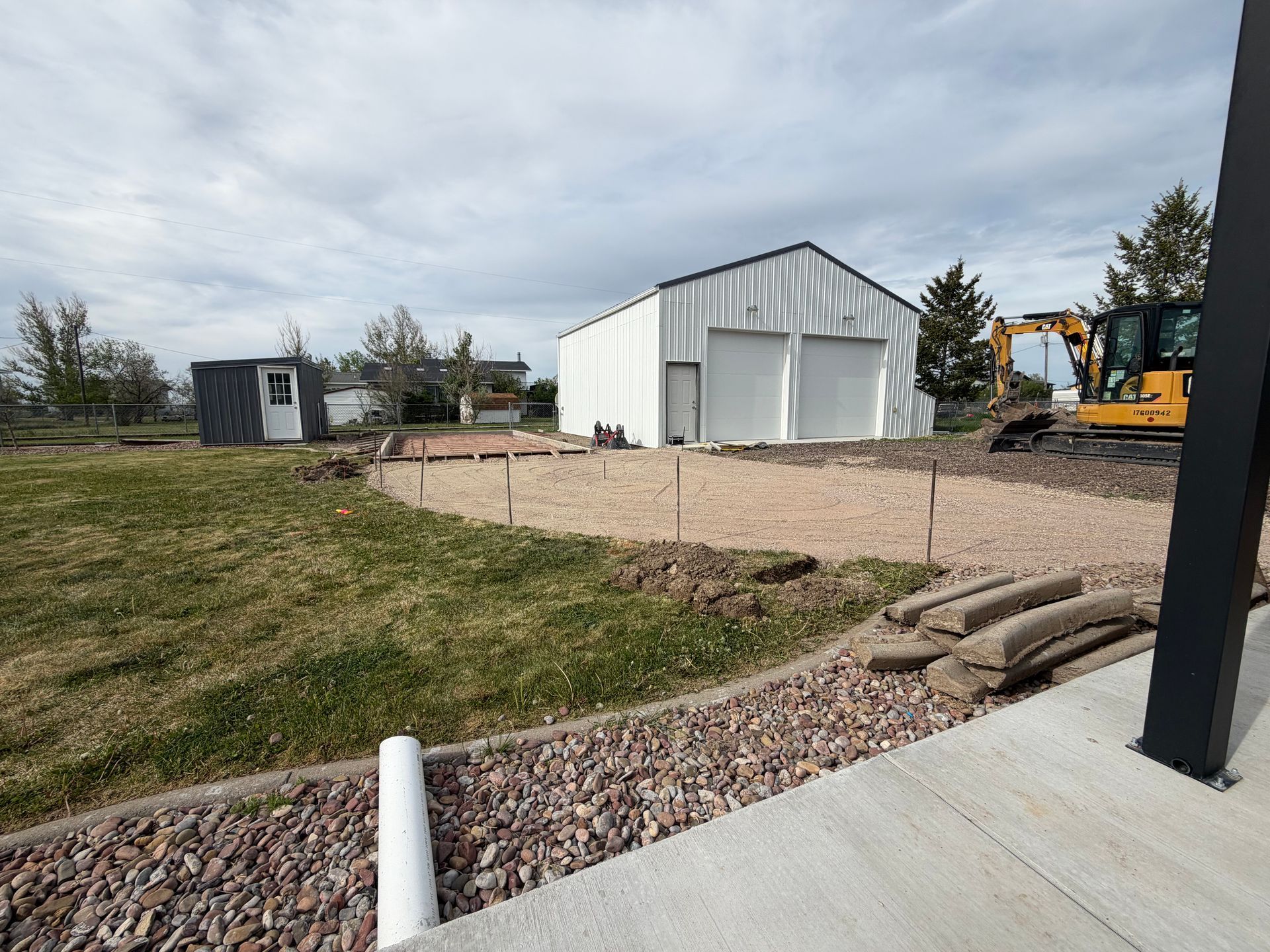 Gravel yard with a white metal shed, small black outbuilding, and patio in the foreground under cloudy sky