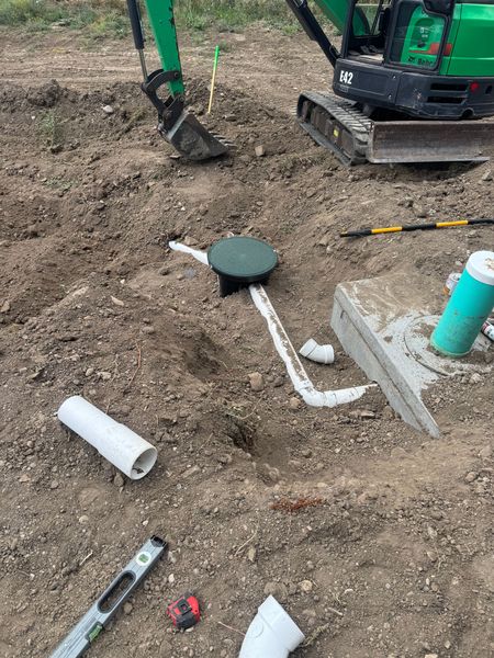 Construction site trench with white PVC pipes and a green utility box in dirt near an excavator track.