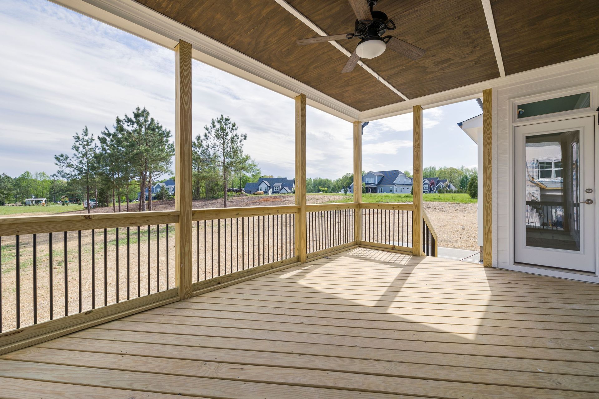 Covered wooden porch overlooking a grassy field, with railings, ceiling fan, and sunlit floorboards.