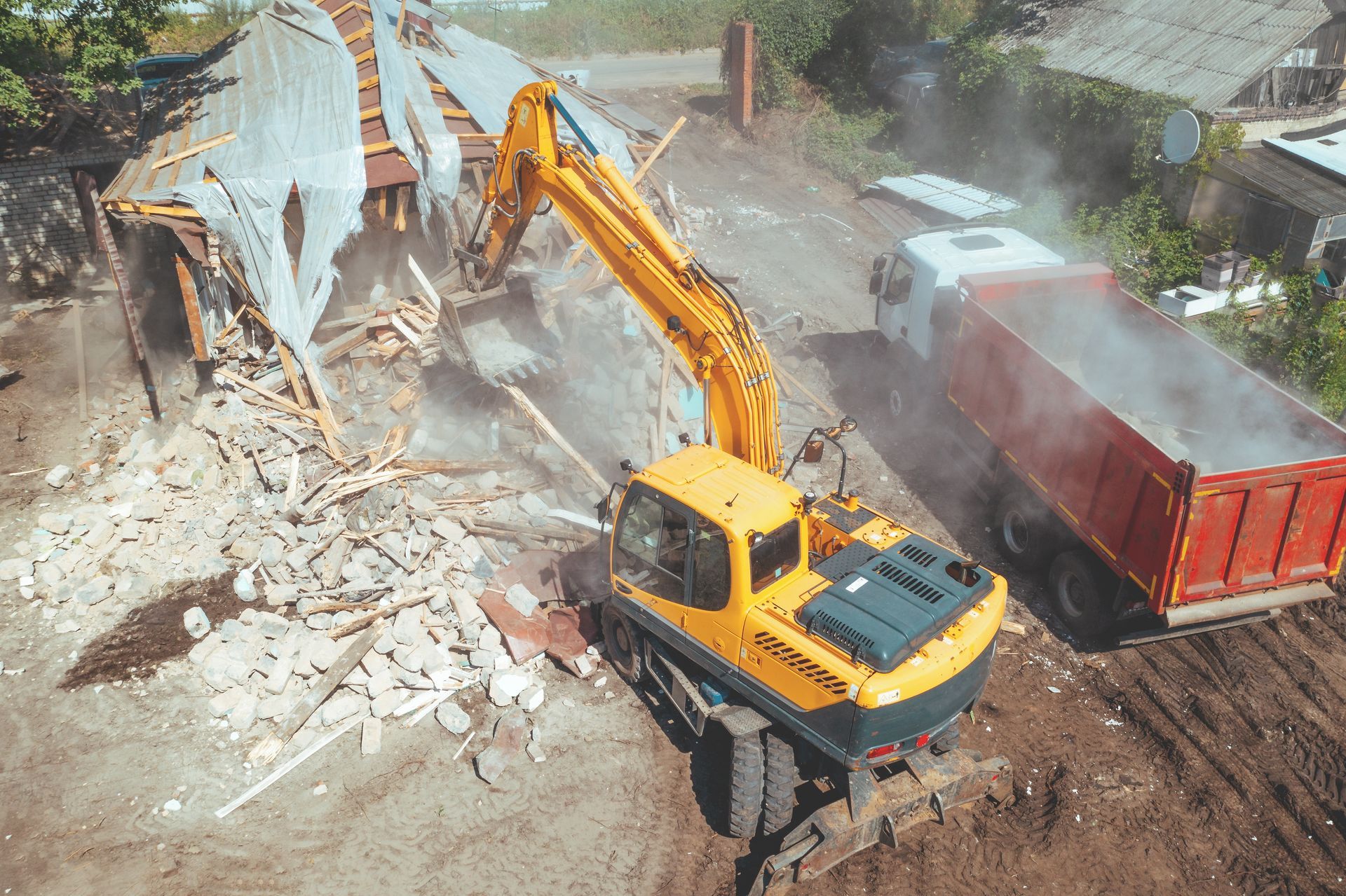Yellow excavator demolishing a building beside a red dump truck amid rubble and dust