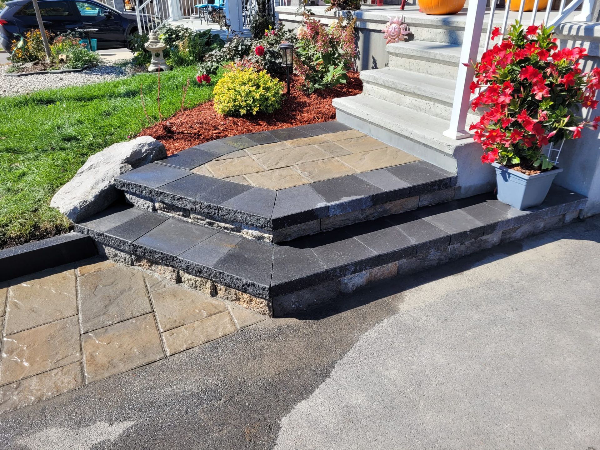 Stone steps leading up to a house with red flowers in a blue pot on the side. Brown and black pavers are used.