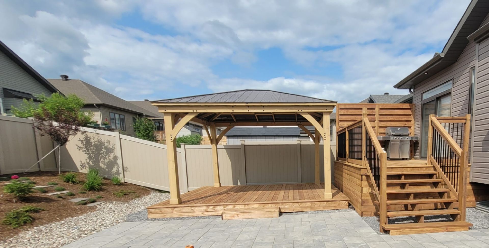 Wooden gazebo on a wooden deck attached to a house, with stairs leading up. A white fence and gray sky are in the background.