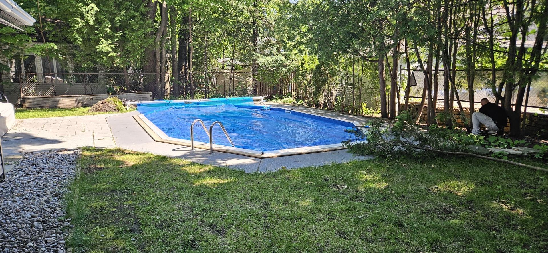 A backyard with a pool covered in blue tarp surrounded by trees and grass. A person is tending to a pile of brush.