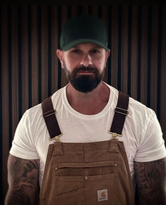 Man wearing green cap, white t-shirt, and brown overalls, with tattoos visible on his arms, in front of a wood-paneled wall.