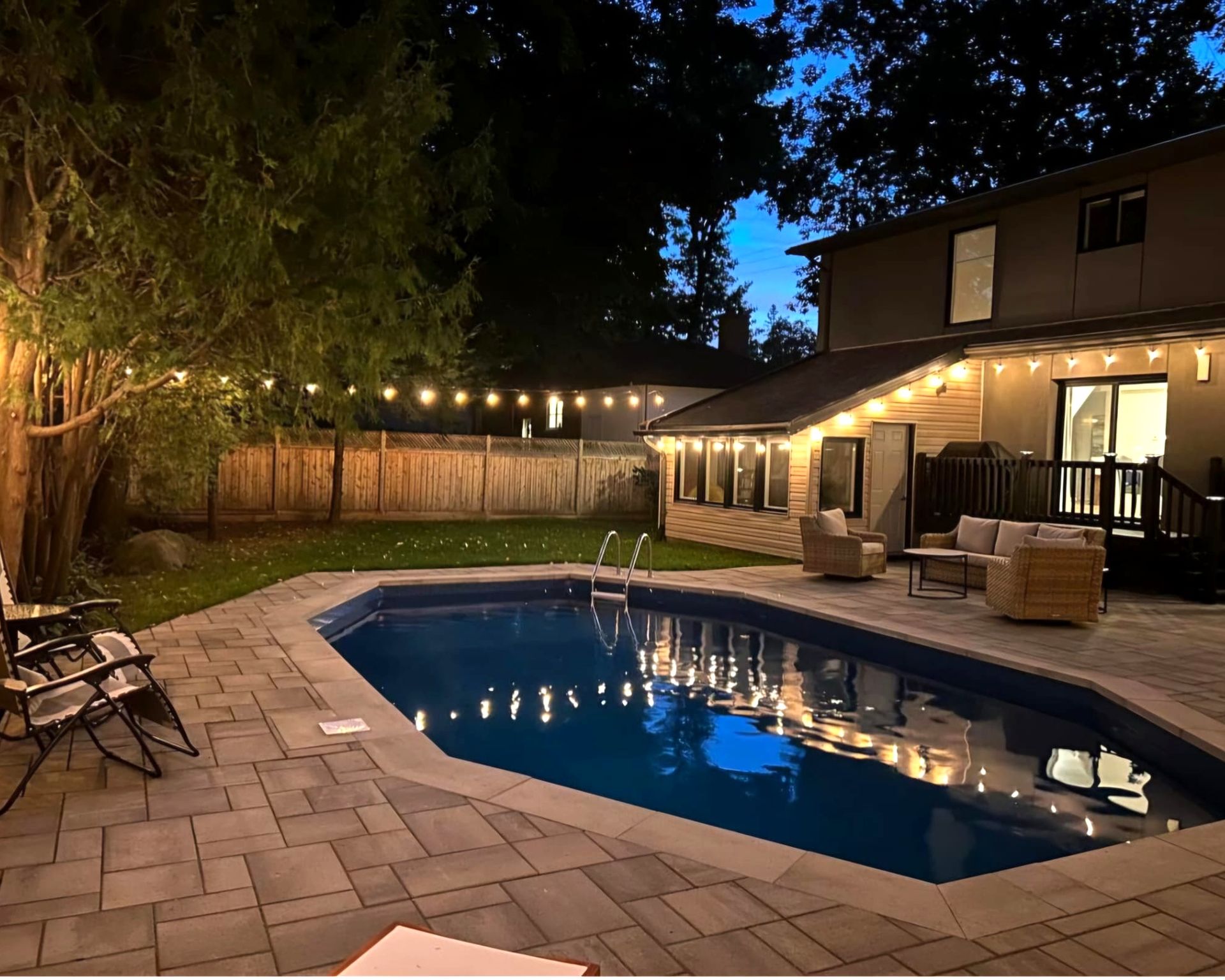 Backyard pool area lit with string lights at dusk. House and wooden fence in background.