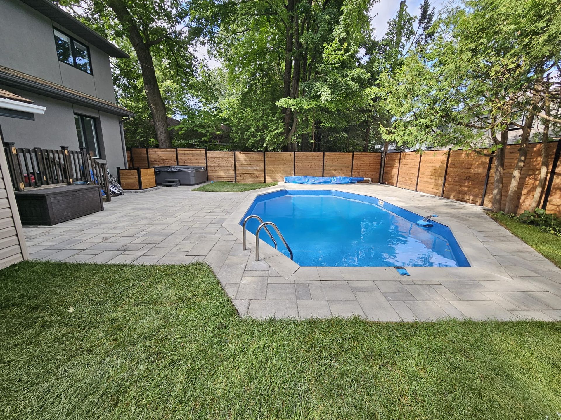 Backyard with a pool surrounded by a paved patio, grass, and a wooden fence. A house is on the left.