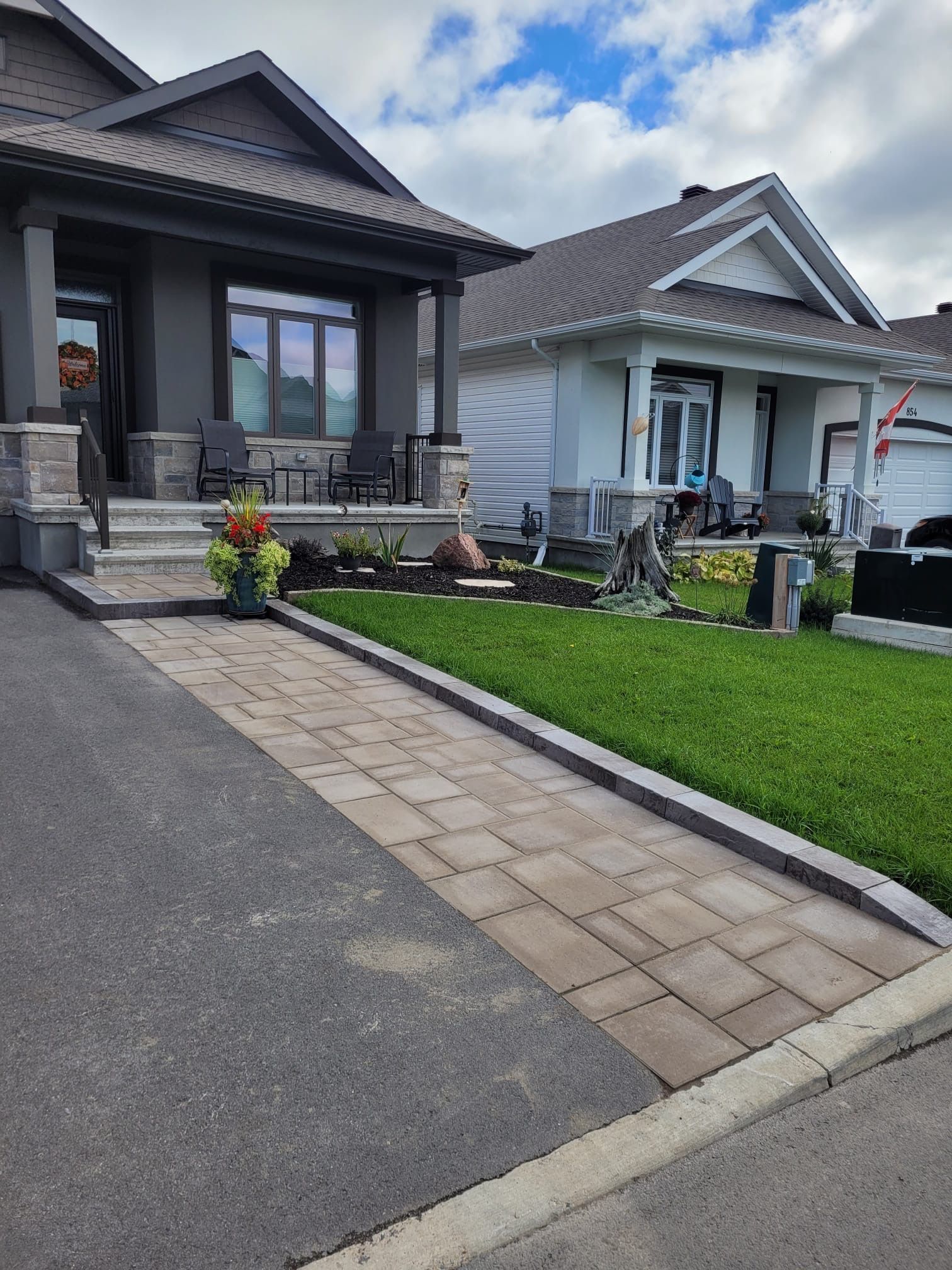 Paved pathway leads to a gray house with a porch. A white house is in the background on a sunny day.