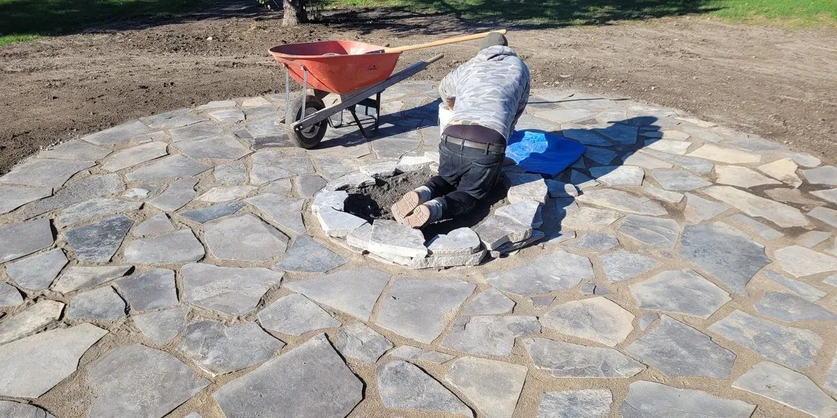 A man is working on a stone patio with a wheelbarrow in the background.