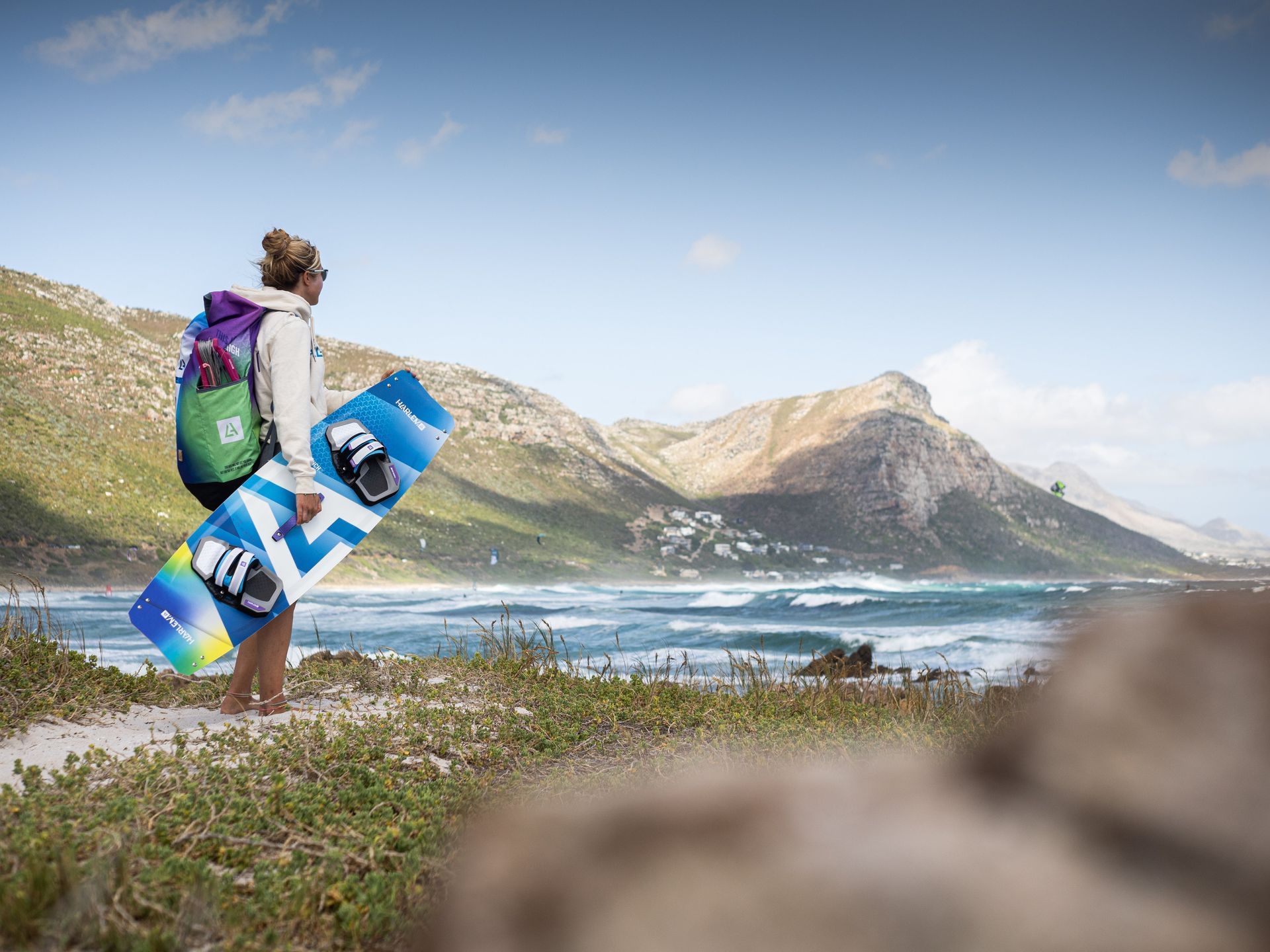 Mujer con mochila y tabla de kitesurf con vistas al océano y las montañas.