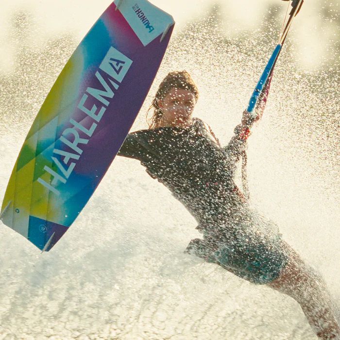 Kiteboarder en el aire con una tabla de colores, con agua salpicando a su alrededor, condiciones soleadas.
