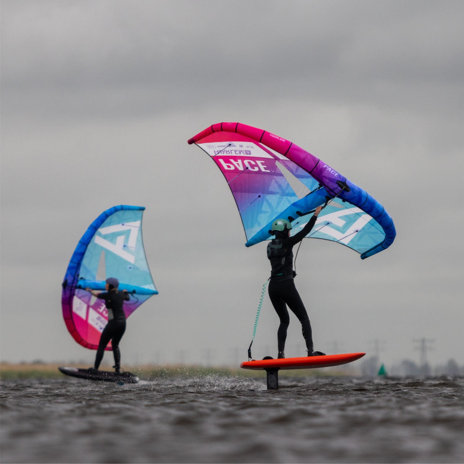 Dos personas practicando wing foil sobre el agua con alas de colores. Cielo nublado.