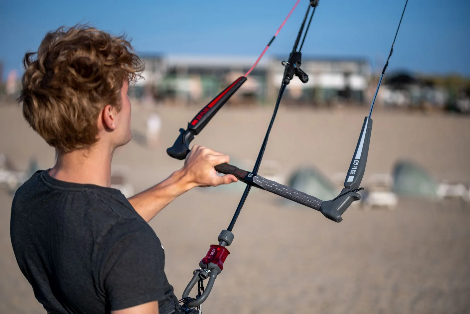 Hombre sosteniendo la barra de control de kitesurf en una playa, cielo azul en el fondo.
