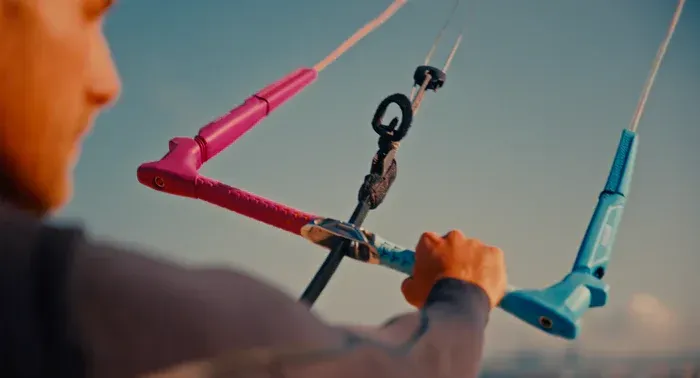 Persona sosteniendo una barra de kitesurf con mangos rosas y azules contra un cielo brillante.